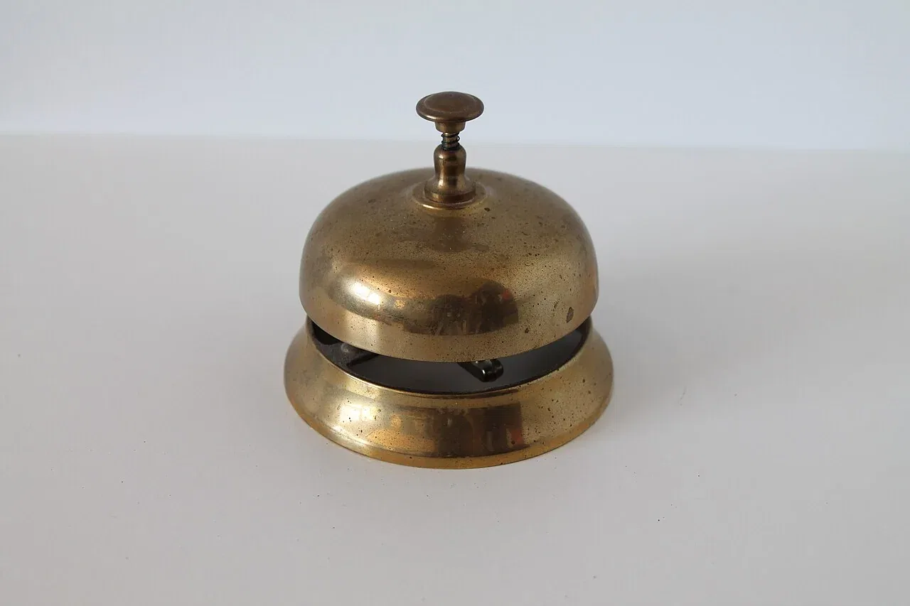 A small brass service bell with a button on top stands on a white surface.