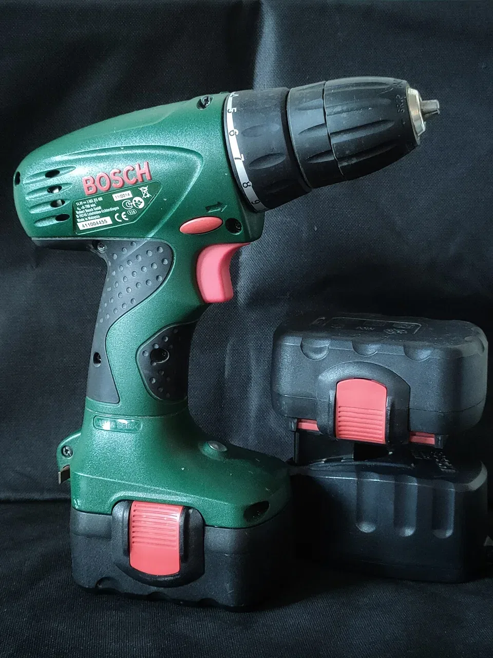 A green cordless drill stands beside a spare battery on a dark cloth.
