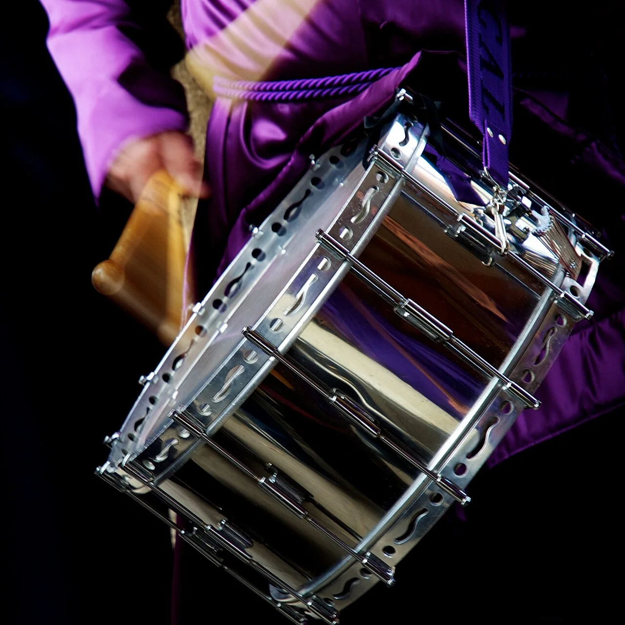 A person in purple robes holds a snare drum with a wooden beater in hand.