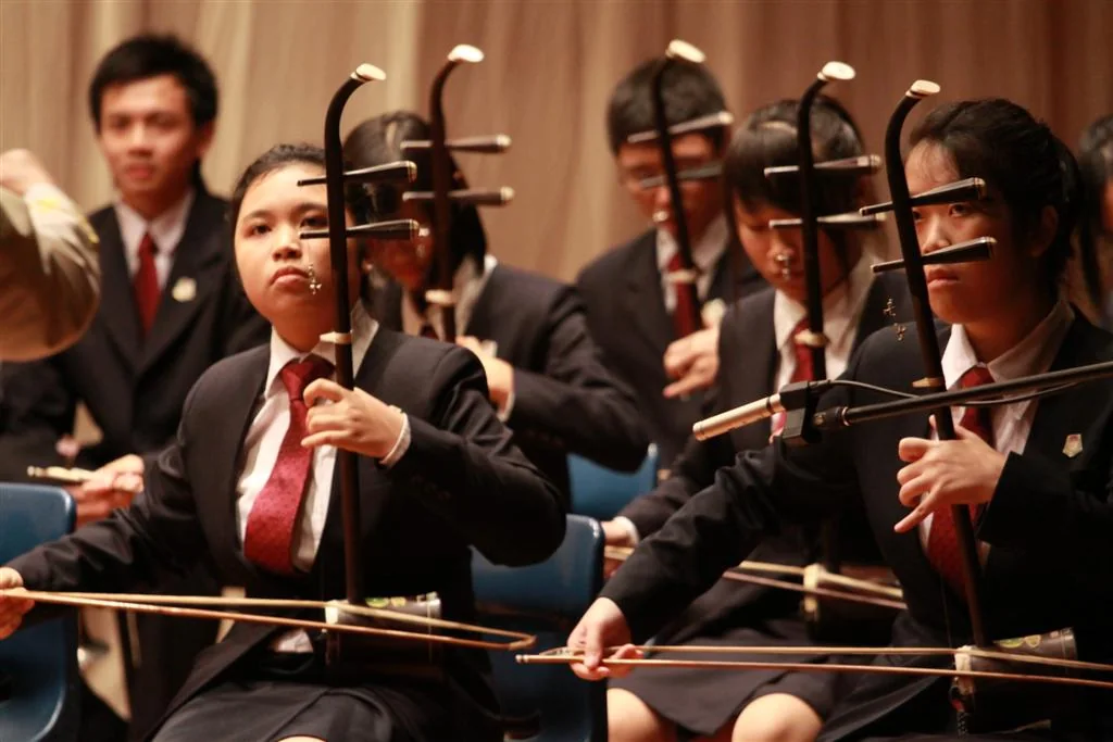 Several musicians in dark suits play erhu instruments while seated in an orchestra.