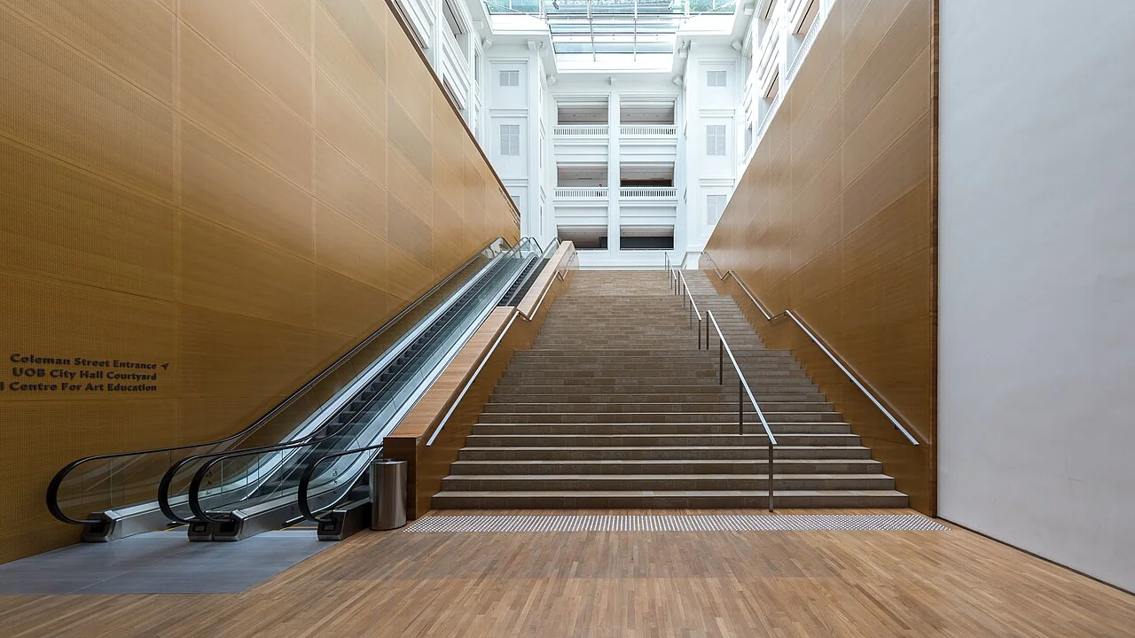 An escalator with glass balustrades runs alongside a wide staircase in a wood-paneled gallery interior.