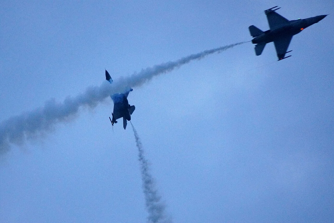 Two F-16 fighter jets fly through a blue sky, one trailing a long white smoke plume.