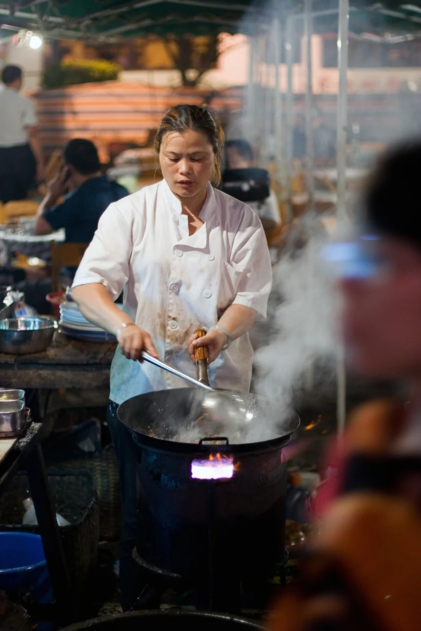 A woman in a white chef coat stirs a wok over a flaming street stove as steam rises around her.