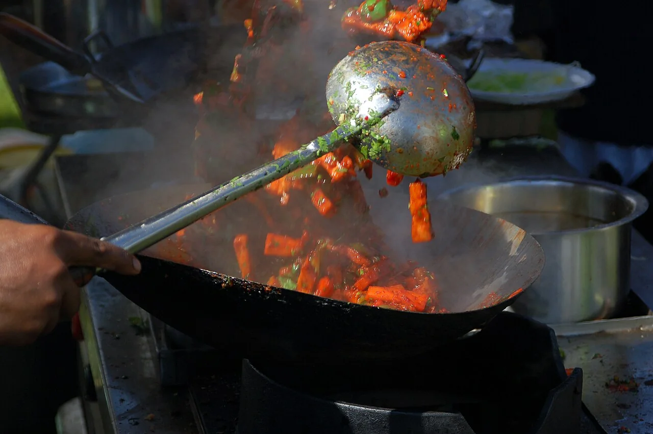 A hand holds a ladle over a wok as pieces of food are tossed amid rising steam.