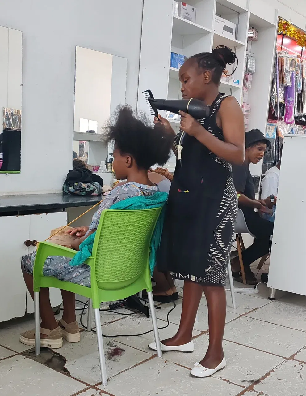 A hairdresser uses a black hair dryer on a seated client's hair in a salon.