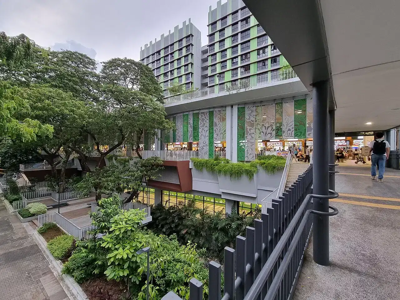 A hawker centre with green floral facade panels sits below residential blocks, seen from a covered walkway.
