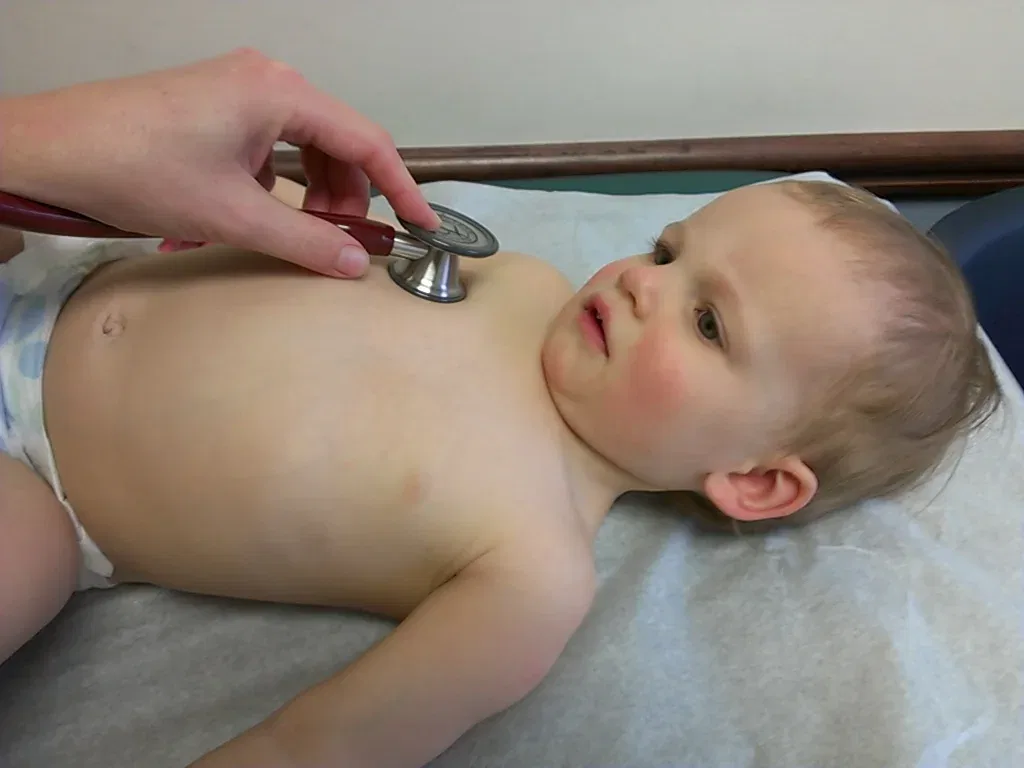 A pediatrician places a stethoscope on the chest of a toddler lying on an examination table.