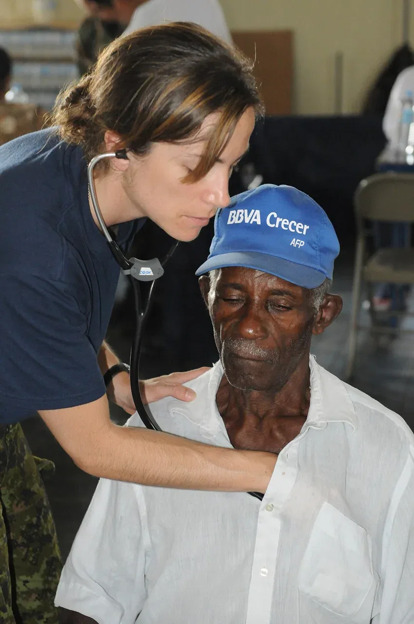 A female medical officer listens with a stethoscope to the chest of an older man wearing a blue cap.