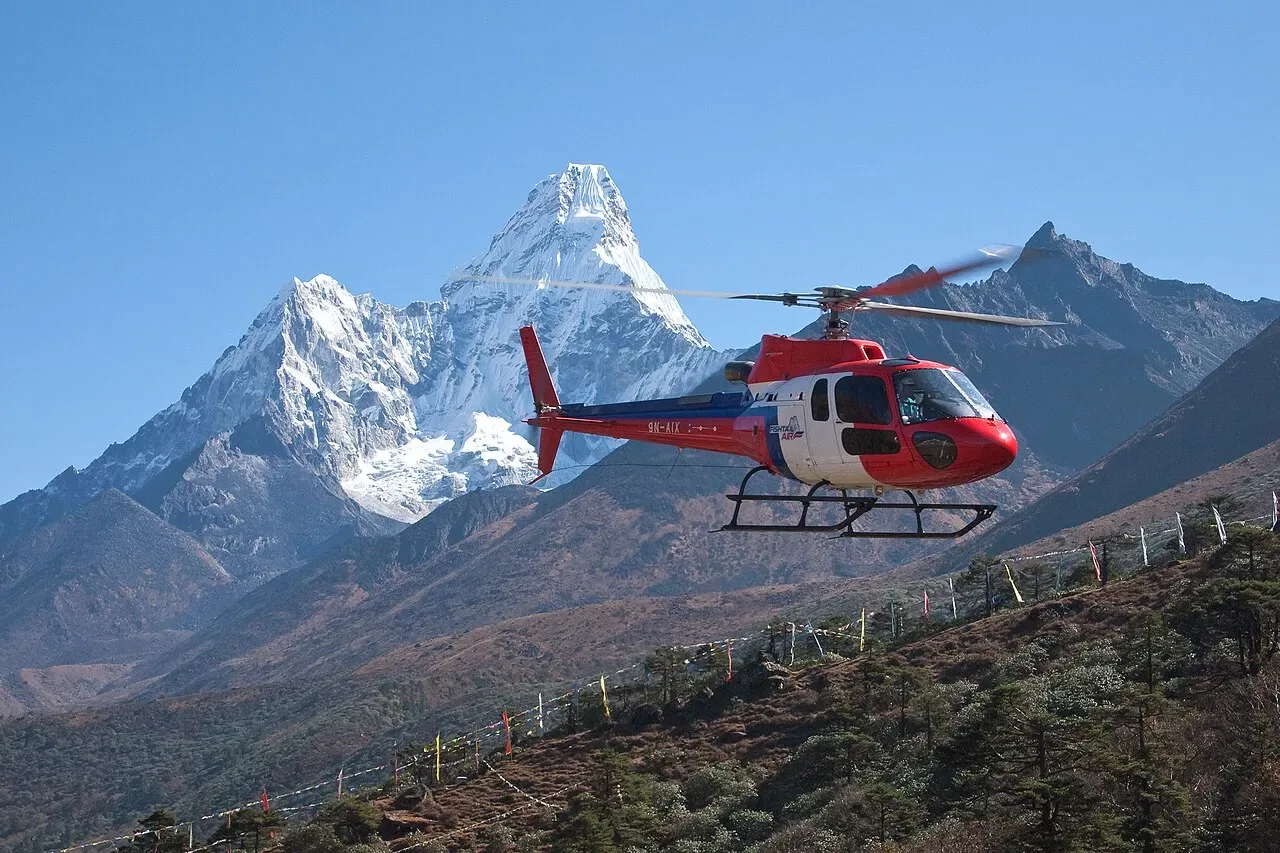 A red and white helicopter flies in front of a snow-capped Himalayan peak under a clear blue sky.