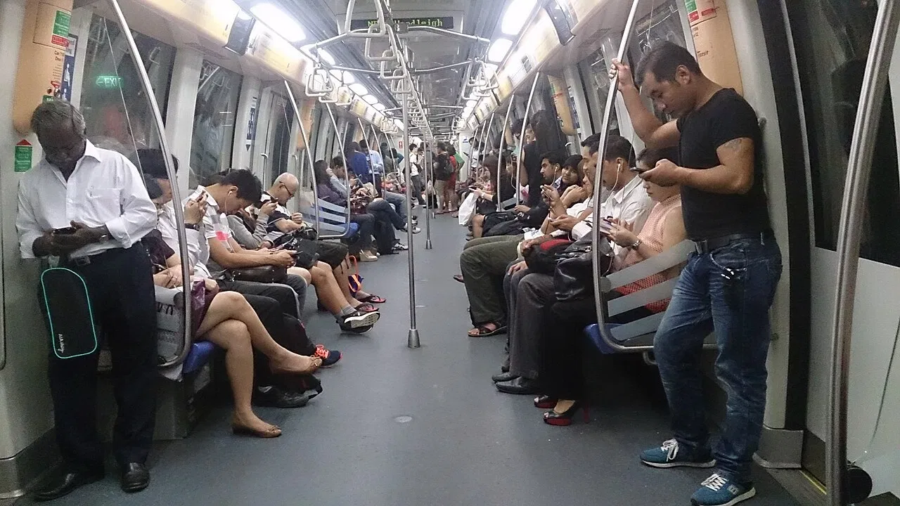 Passengers sit and stand inside a brightly lit Singapore MRT carriage, most looking at their phones.