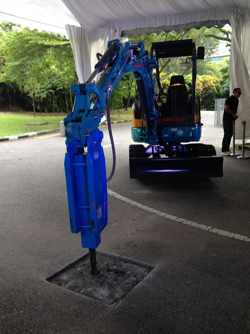 A blue hydraulic jackhammer mounted on a small excavator rests its chisel over a square opening in the asphalt.