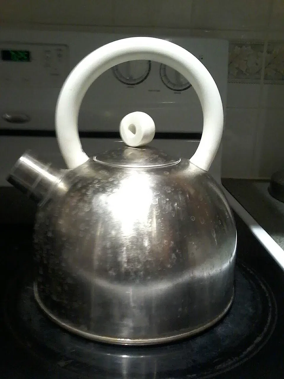 A stainless steel whistling kettle with a white handle sits on a black stovetop.