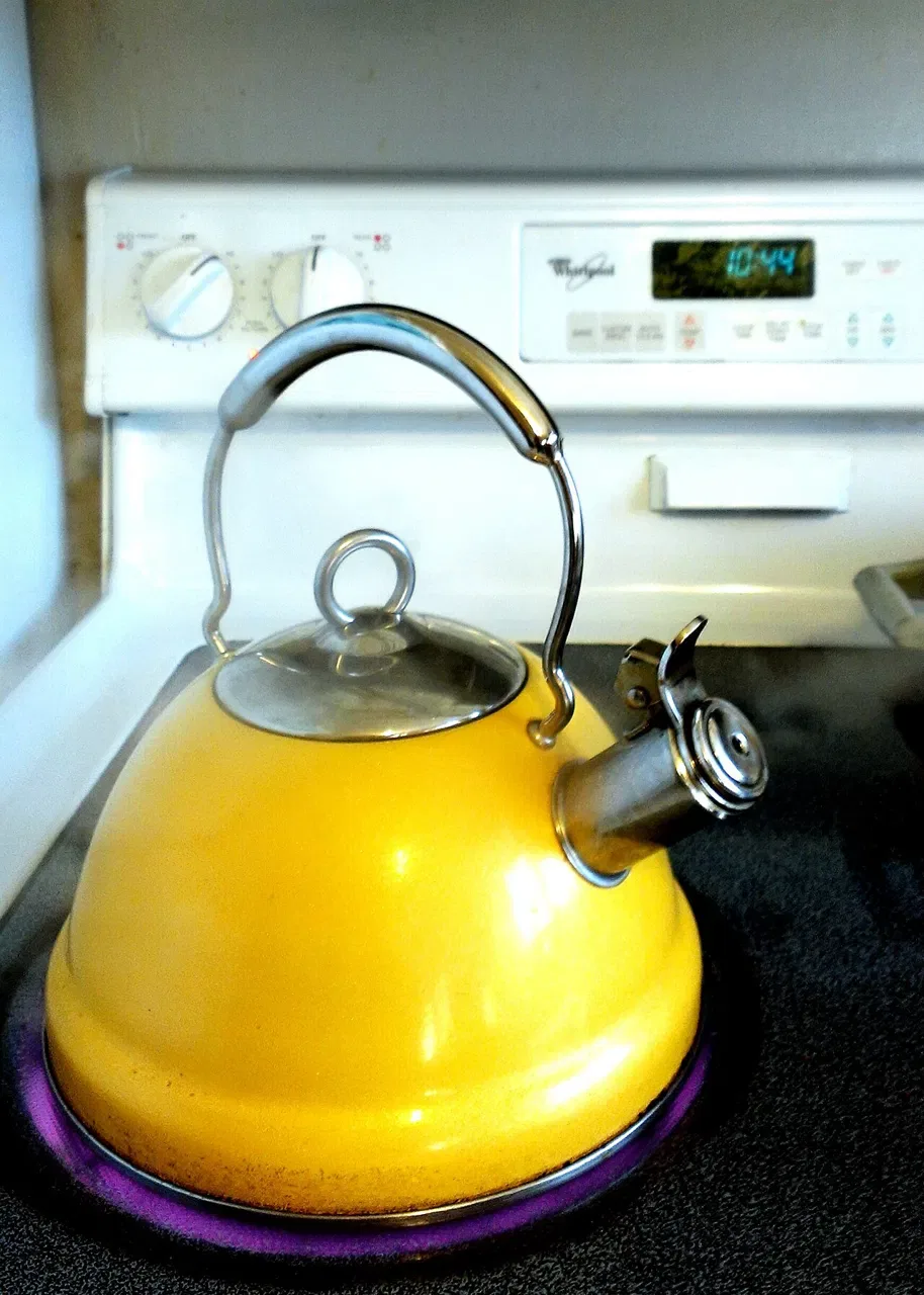 A yellow whistling kettle sits on a black electric stovetop with steam escaping from its spout.