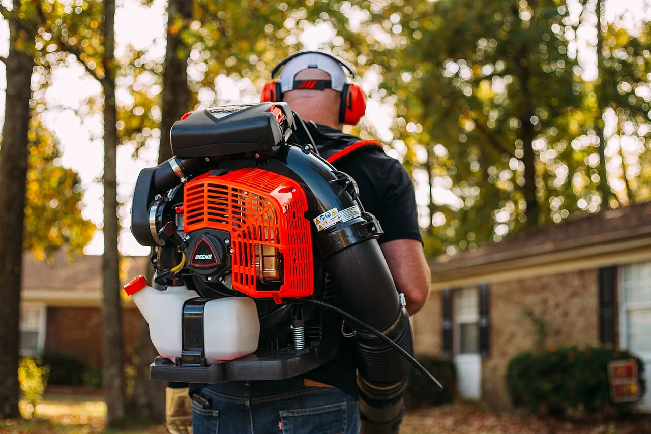 A man wearing a backpack leaf blower stands outdoors near trees and a house.
