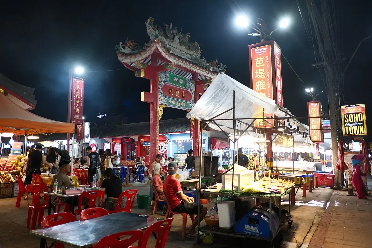 Red plastic tables and food stalls fill a night market street beneath a decorative Chinese-style gate.