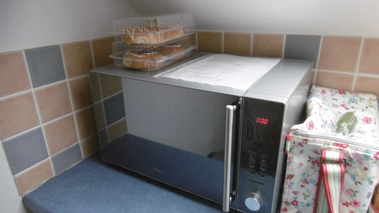 A silver microwave oven sits on a blue counter between tiled walls, with pastries in plastic containers on top.