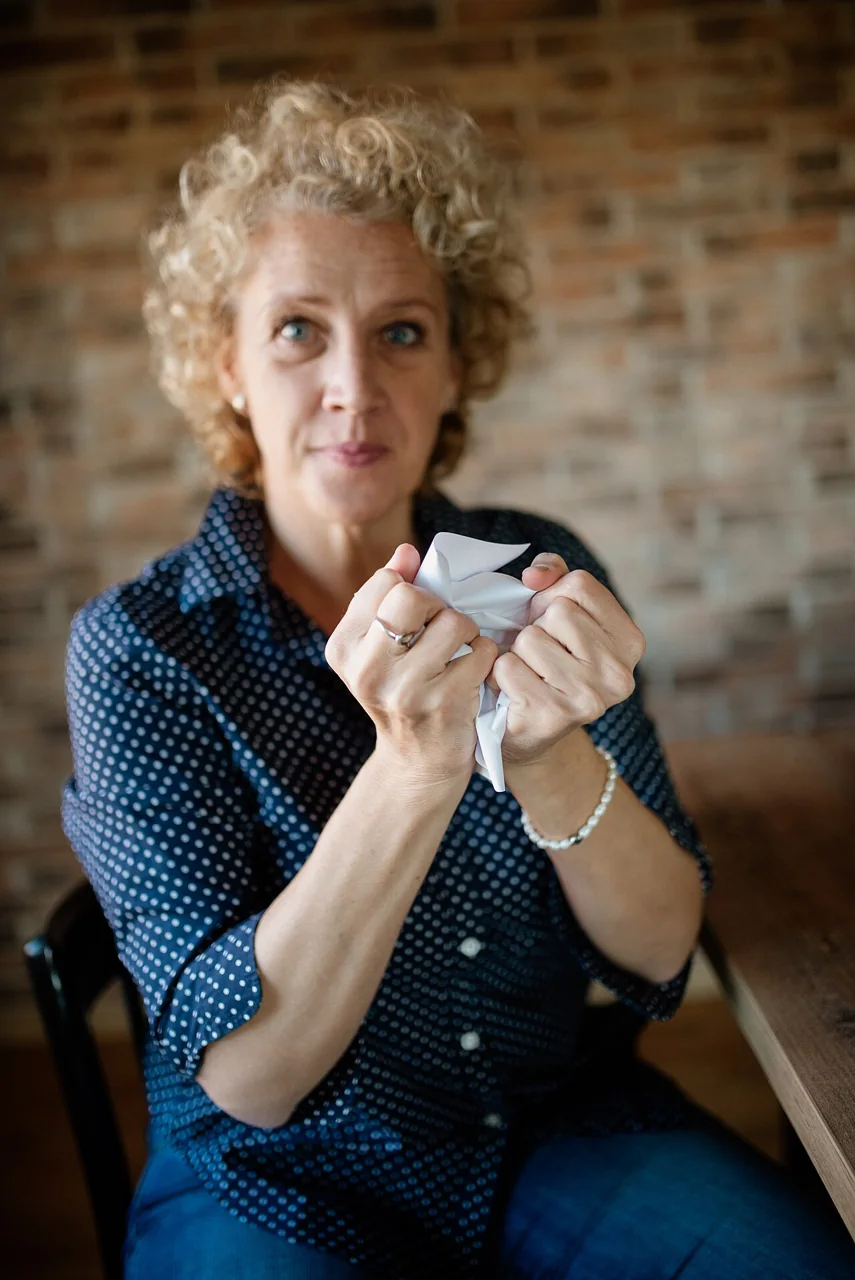A woman in a blue polka-dot shirt holds and crumples a piece of white paper with both hands.