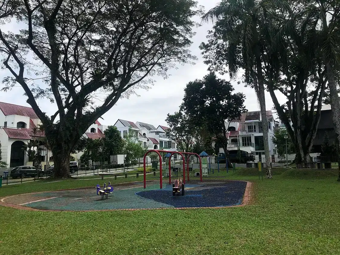 Red swing frames and spring riders stand on a blue playground surface beside grass and trees.