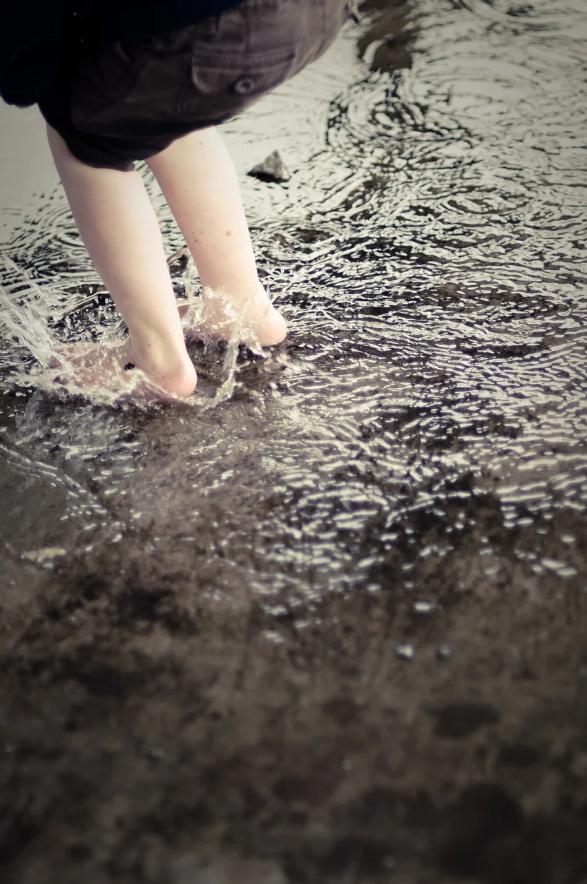 A child's bare feet splash water while stepping in a shallow puddle.