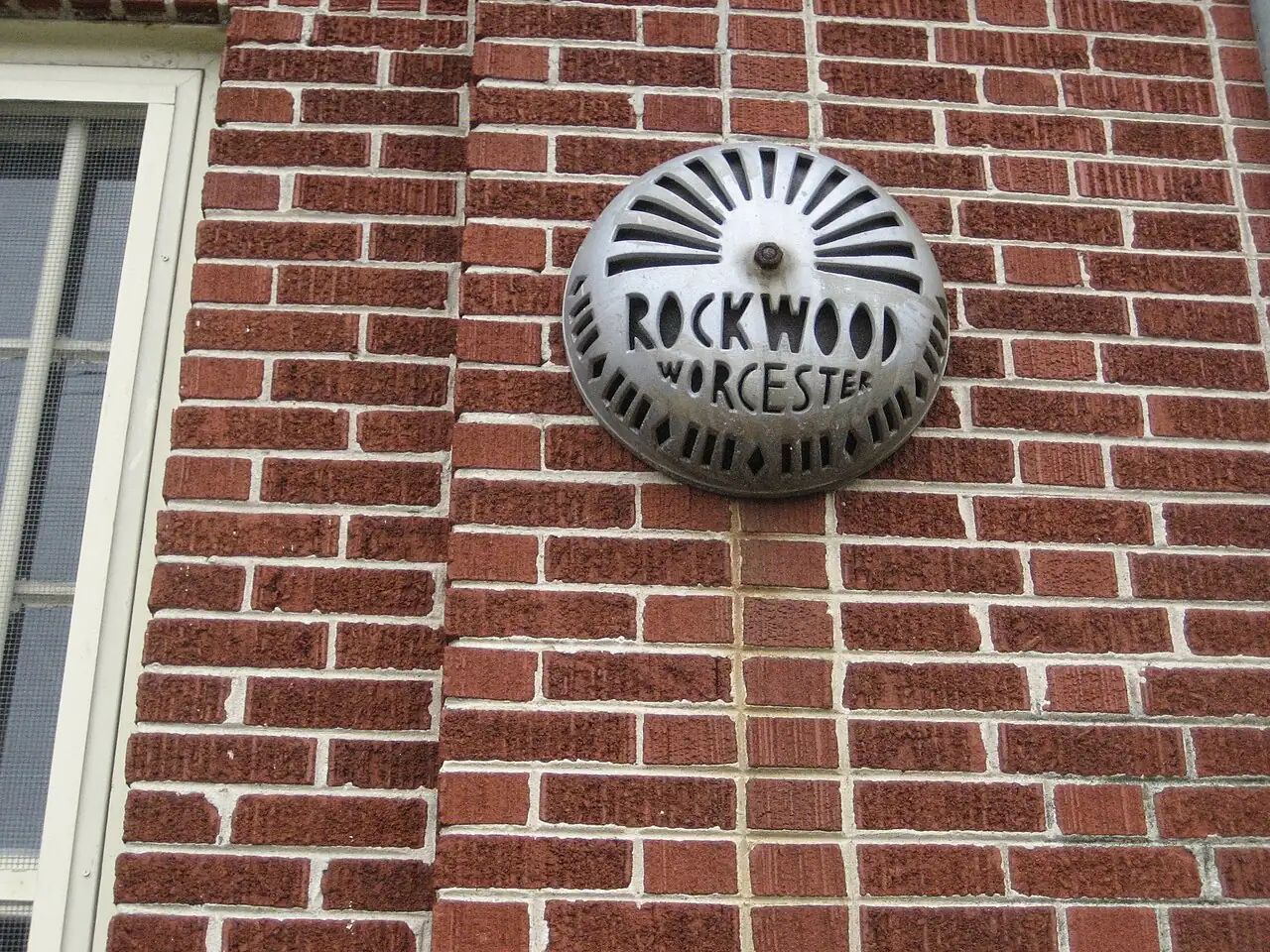 A round metal bell marked ROCKWOOD WORCESTER is mounted on a red brick wall beside a window.