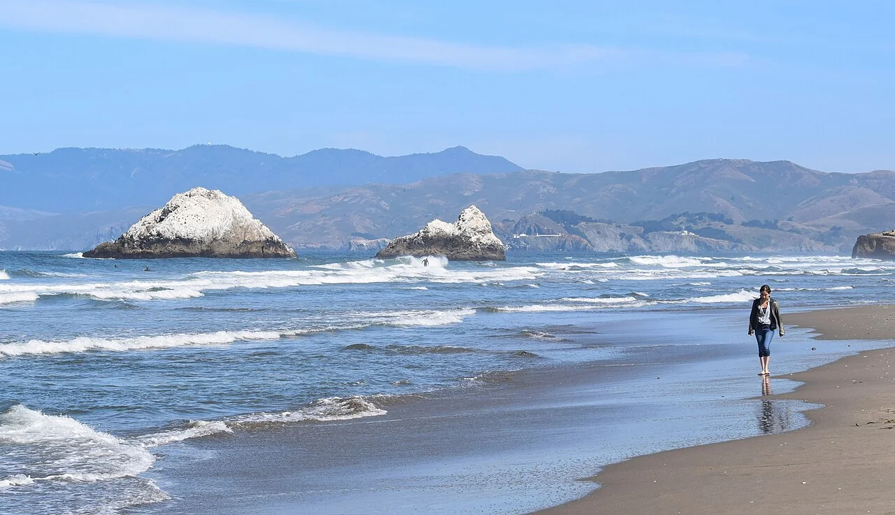 Waves break on a sandy beach with a person walking along the wet sand and rocky outcrops in the distance.