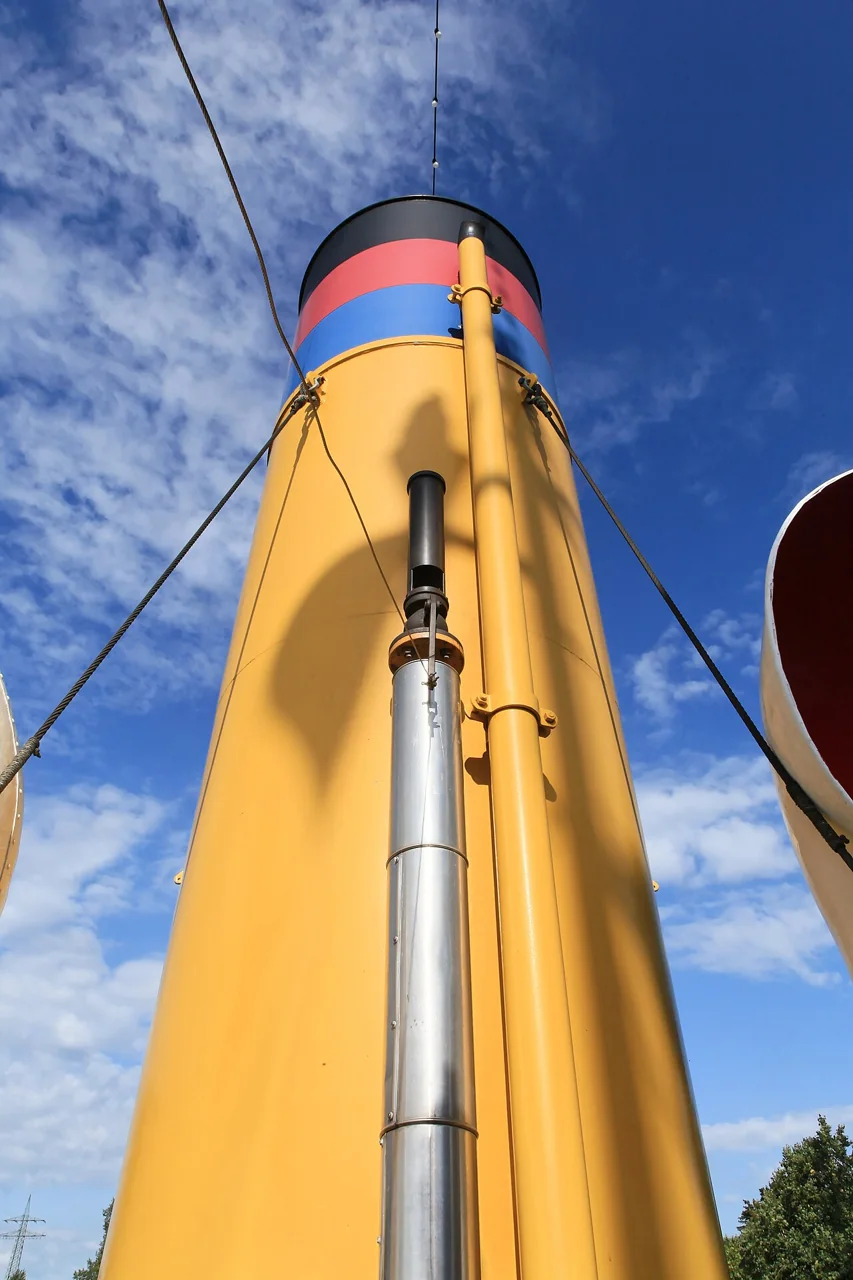 A yellow ship funnel with black, red and blue bands rises against a blue sky, with a silver steam whistle pipe attached to its side.