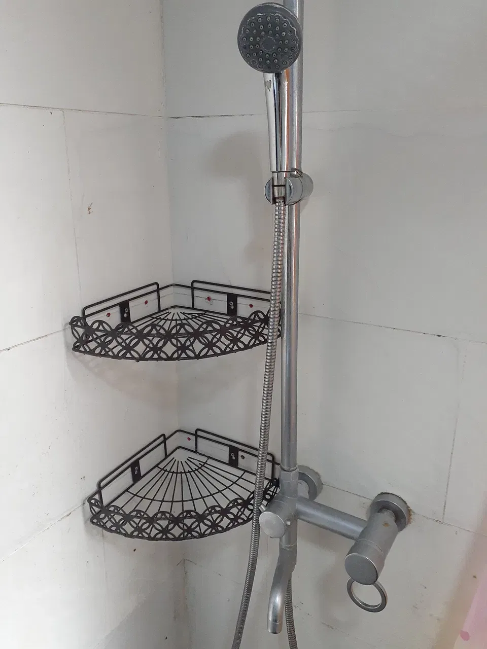 A handheld showerhead with a metal hose hangs above two black wire corner shelves on a tiled wall.