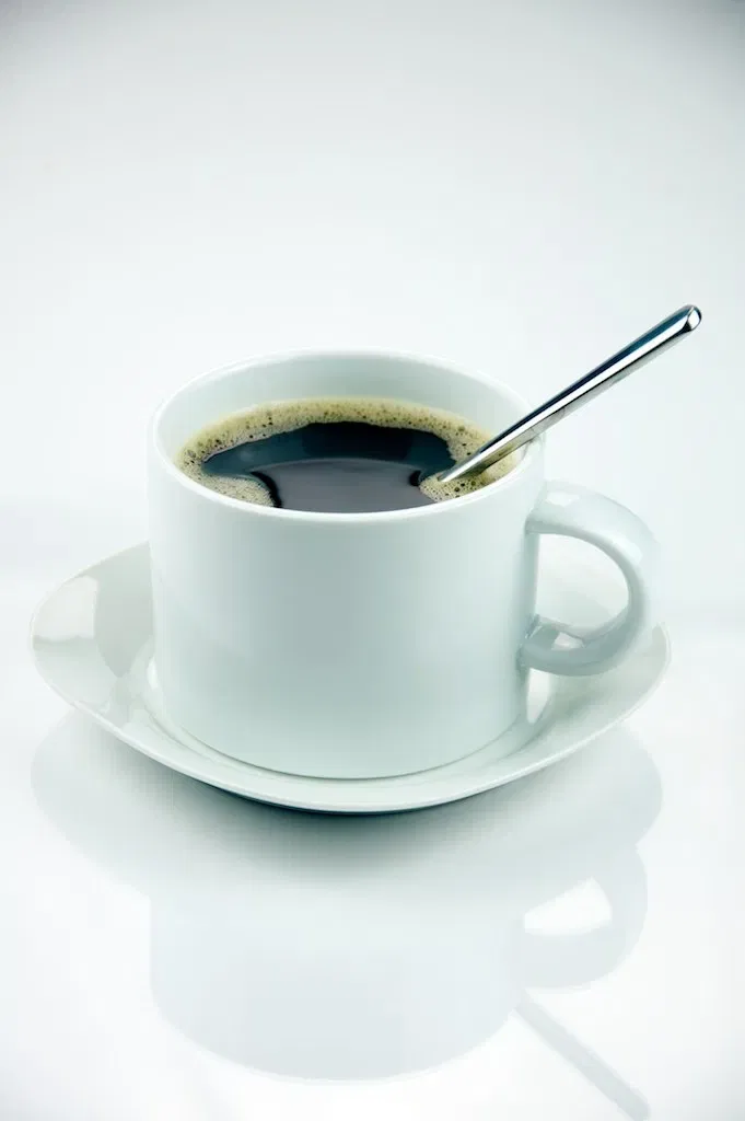 A metal spoon rests in a white cup of black coffee on a saucer.