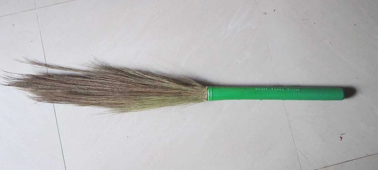 A broom with a green handle and natural bristles lies on a tiled floor.