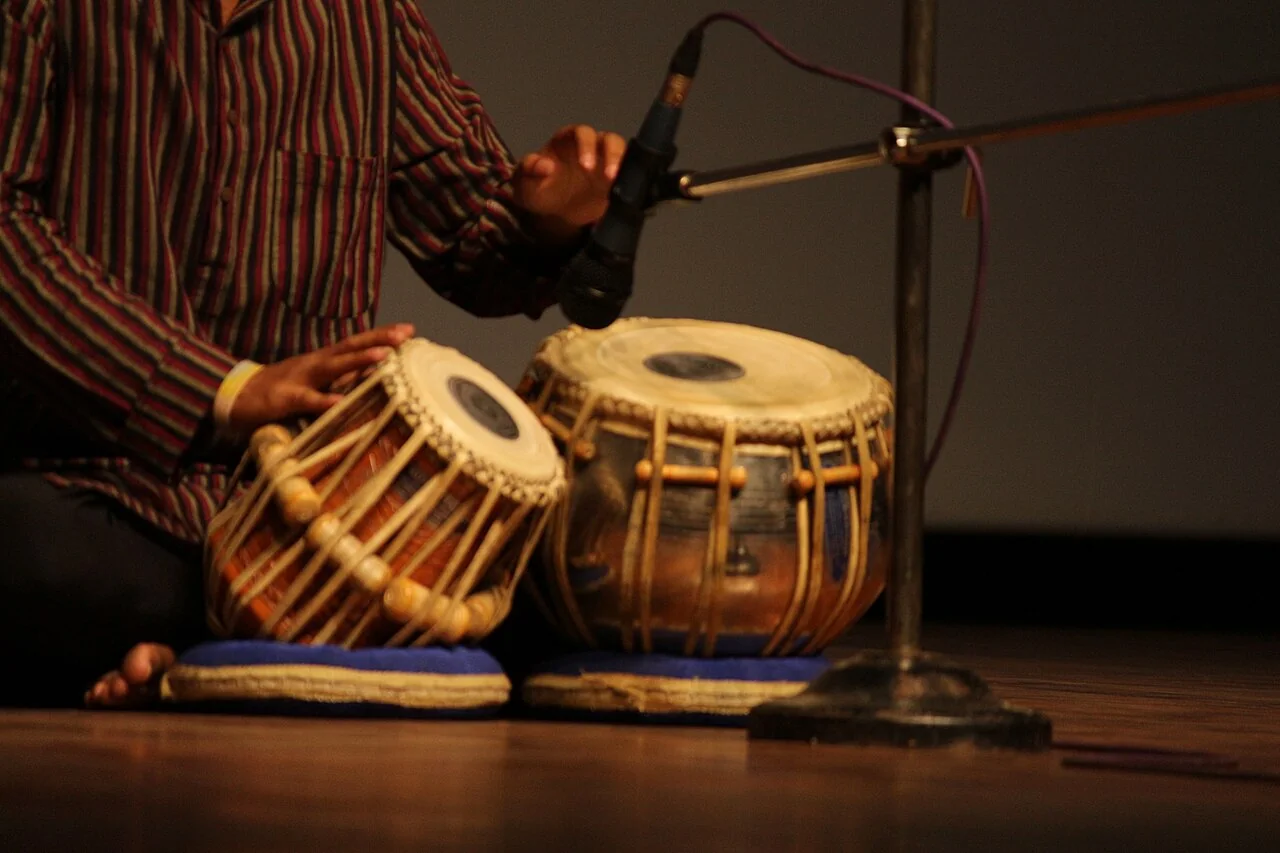 A seated musician holds one hand over a pair of tabla drums beside a microphone.