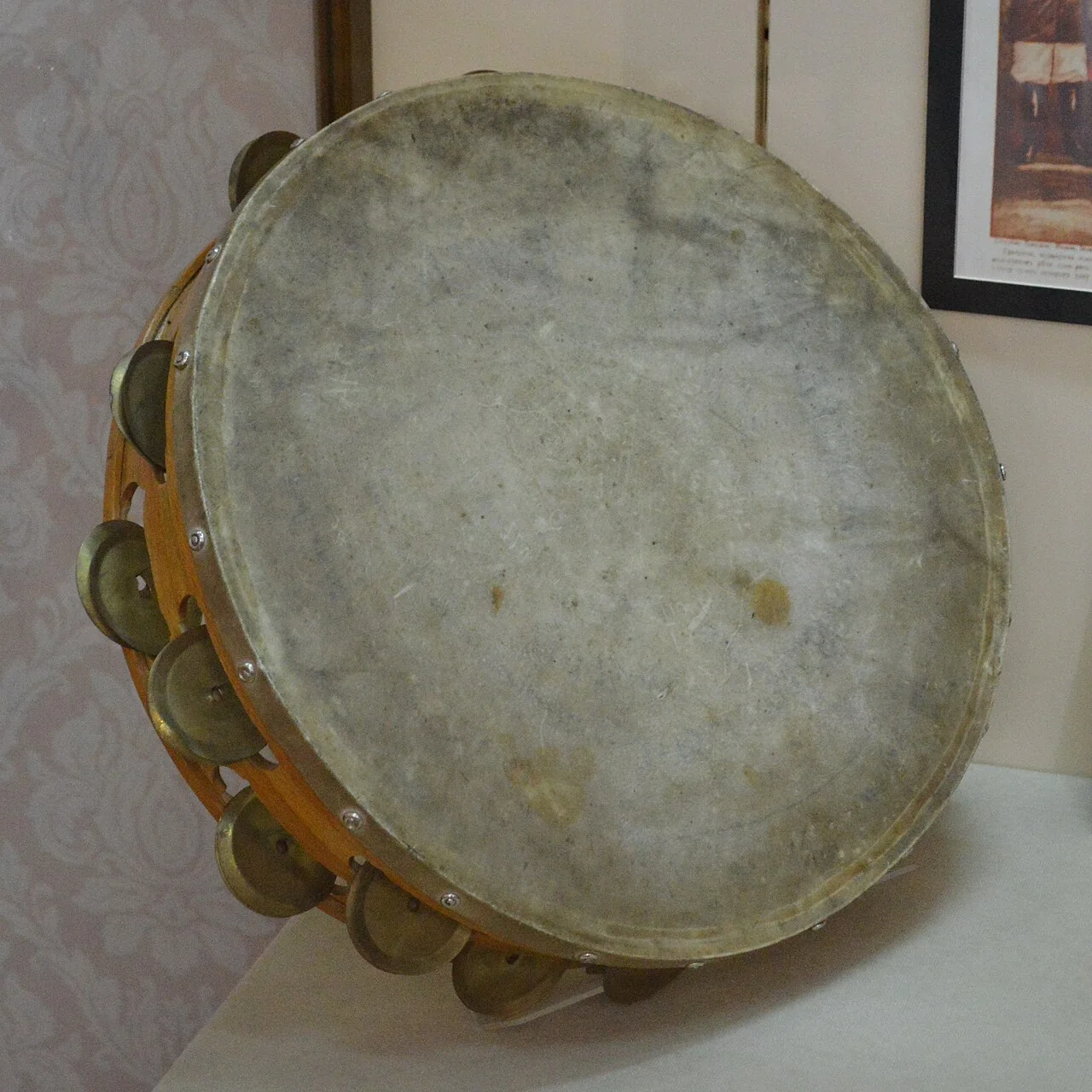 A tambourine with a wooden frame, metal jingles and a worn skin head rests on a white surface.