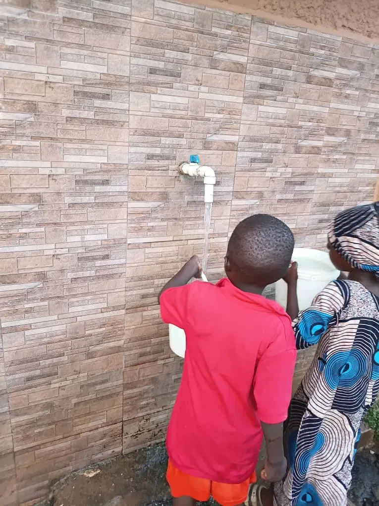 Children collect water from an outdoor tap beside a tiled wall.
