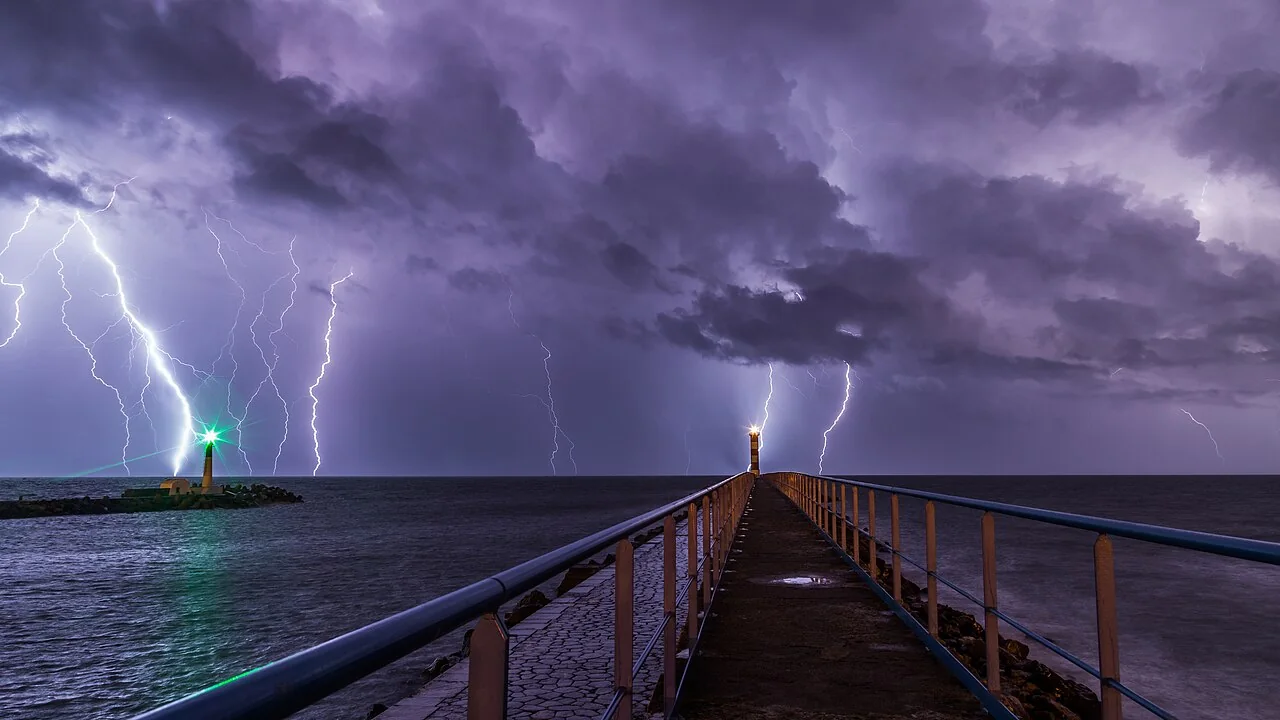Several lightning bolts branch down from purple storm clouds over the sea, with a lit lighthouse on a breakwater and another at the end of a pier.