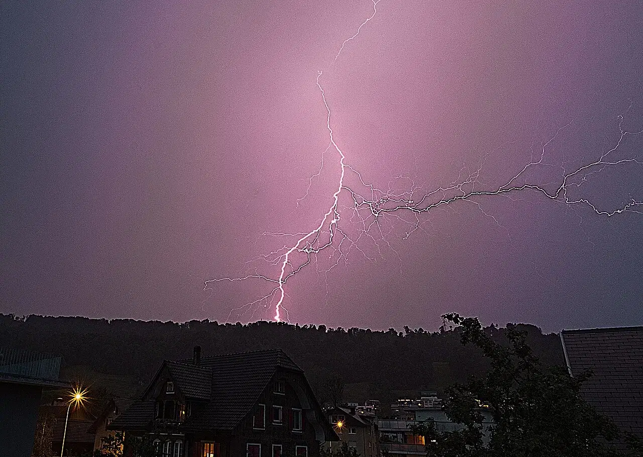 A bright branched lightning bolt splits a purple twilight sky above dark rooftops and a wooded hill.