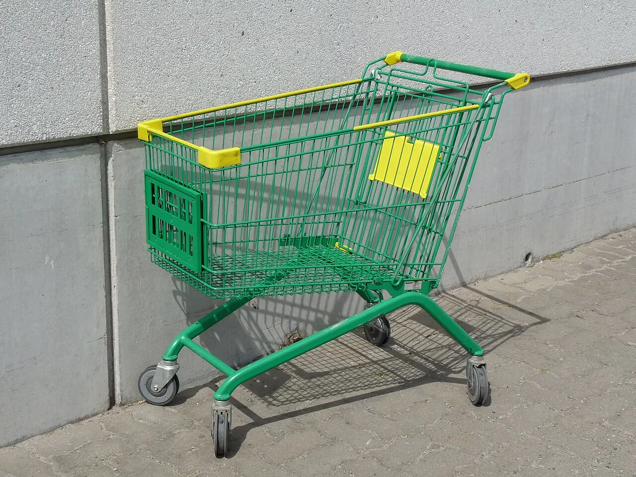 An empty green shopping trolley stands beside a wall.