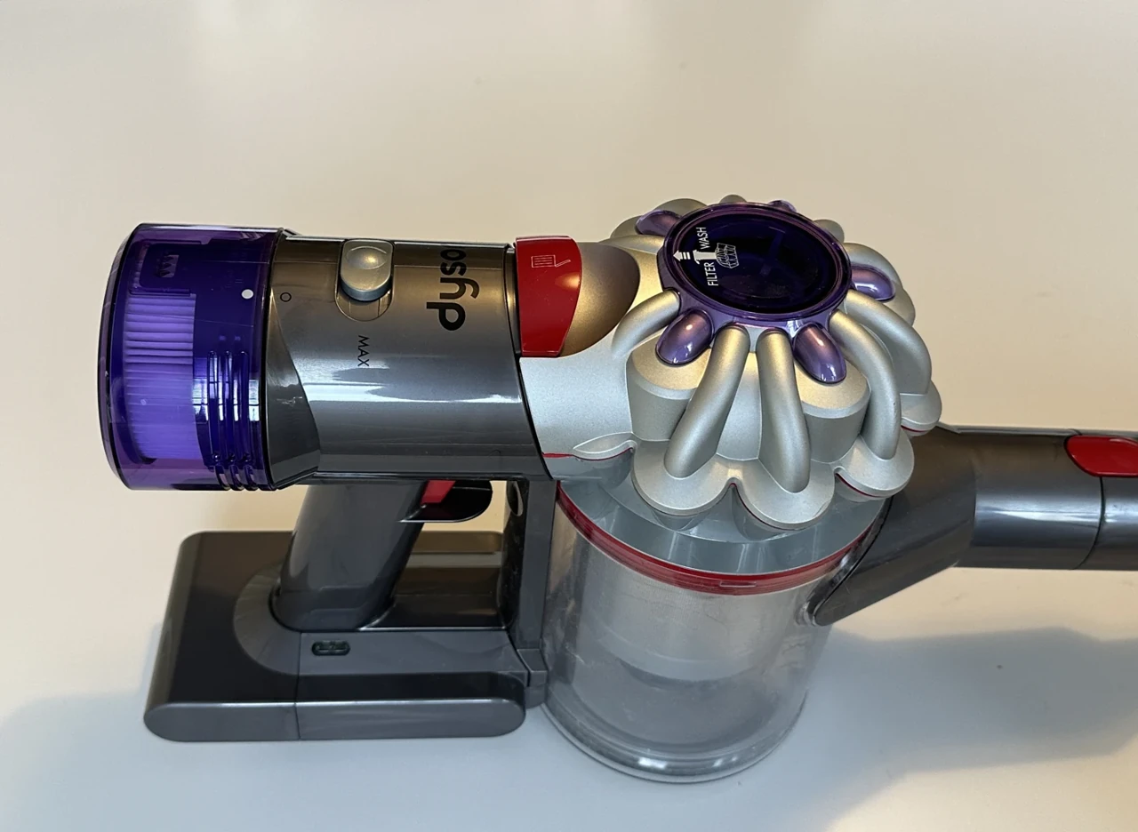 A cordless vacuum cleaner has a grey handle, a clear dust container and purple filters.