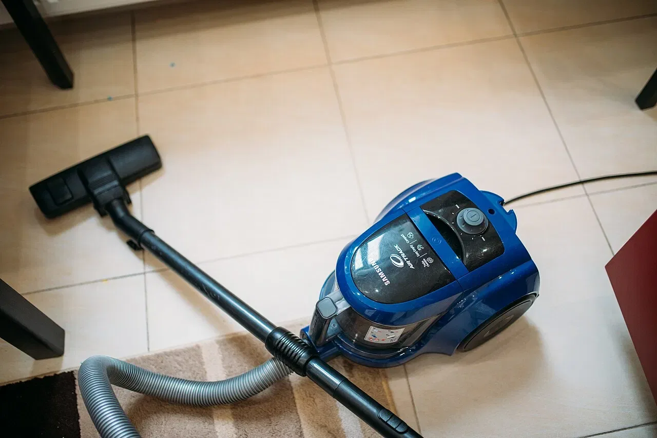 A blue canister vacuum cleaner with its hose and floor nozzle rests on a tiled floor beside a rug.