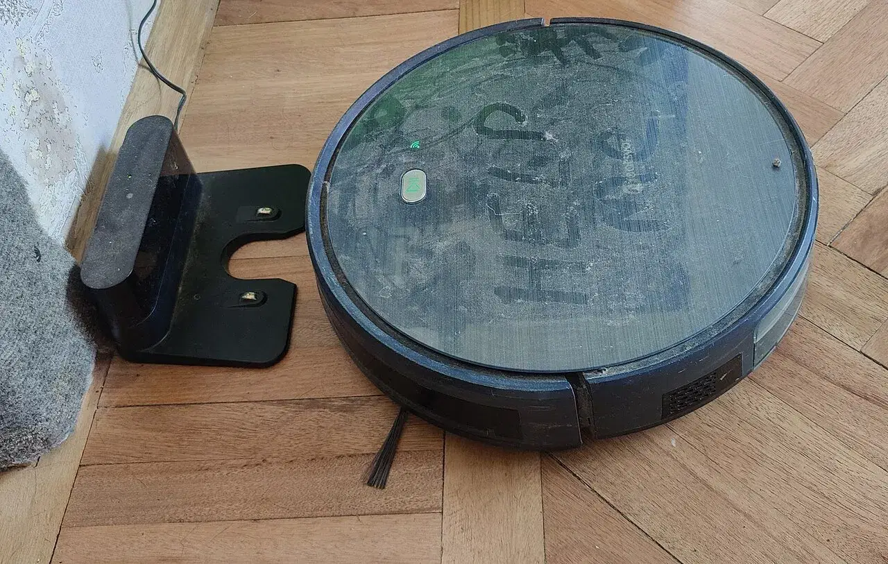 A round robot vacuum cleaner sits on a wooden floor next to its charging dock.