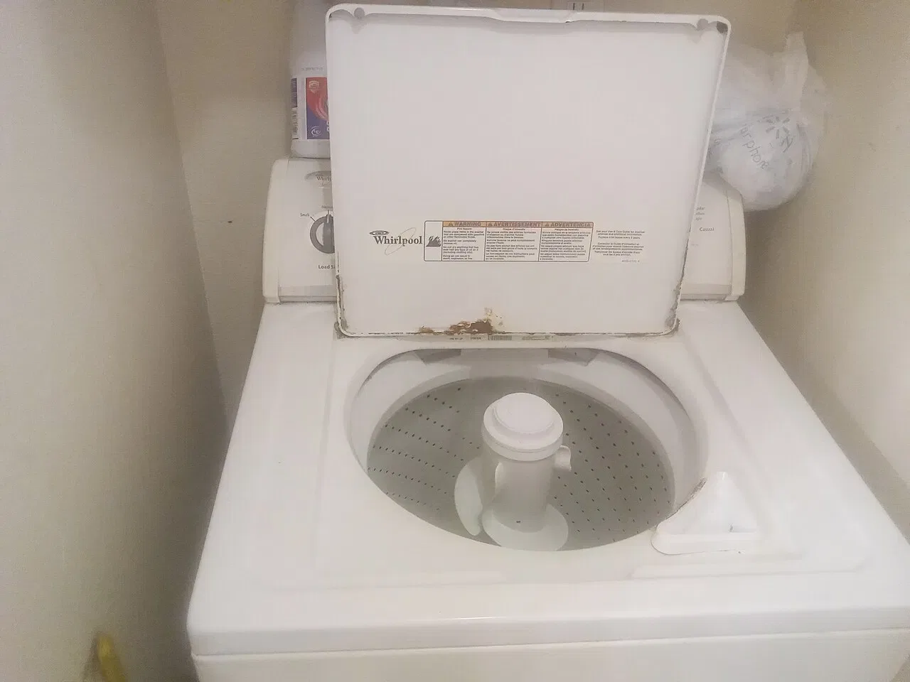 An open top-loading washing machine with an empty drum stands against a wall.