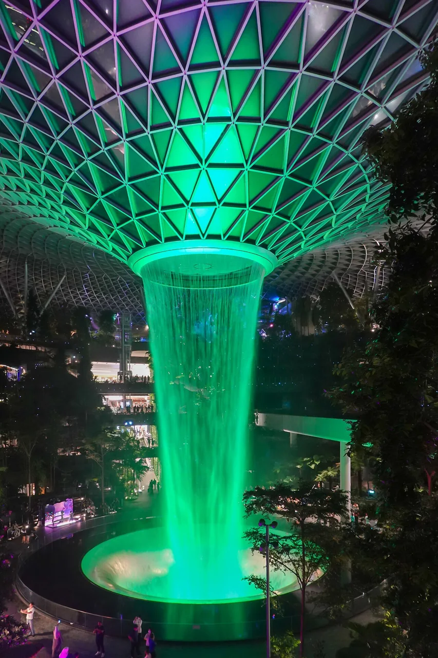 A tall indoor waterfall of green-lit water falls from a ring in a glass-roofed atrium.