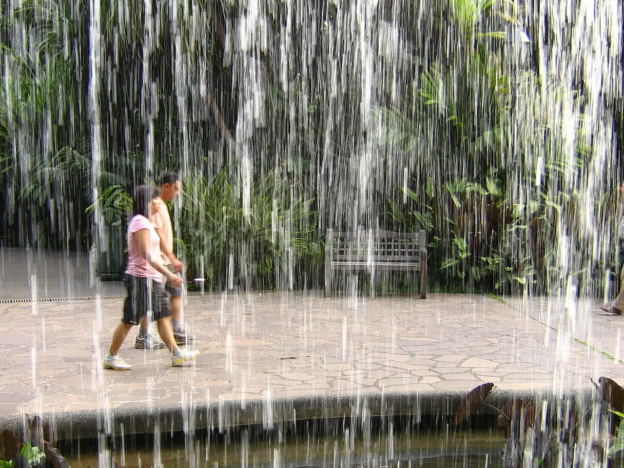 Two people on a paved path are seen through a curtain of falling water.