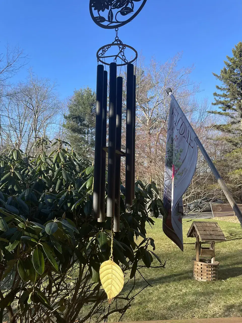 A metal wind chime with black tubes hangs in front of a leafy bush under a blue sky.