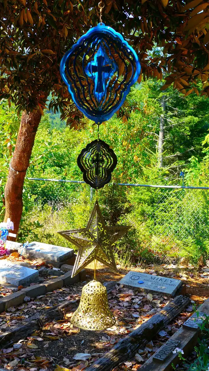 A blue and gold wind chime hangs from a tree branch above a cemetery grave.