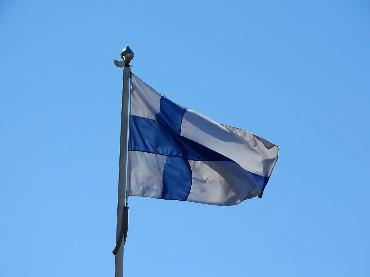A blue-and-white Finnish flag ripples on a pole against a clear blue sky.
