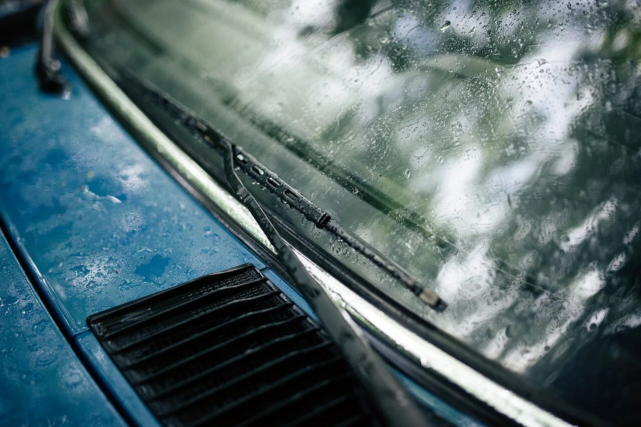 A windshield wiper lies across the rain-covered glass of a blue car.