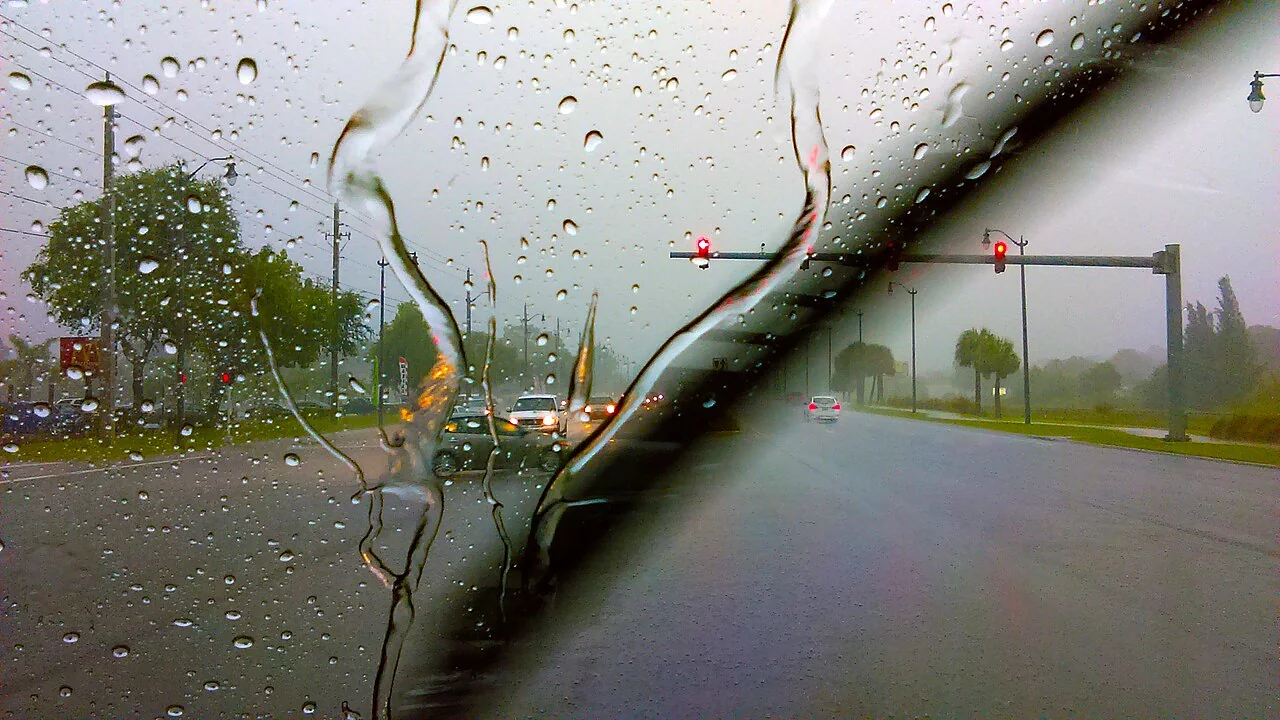 A windshield wiper blade sweeps across a rain-spattered car window with traffic ahead.