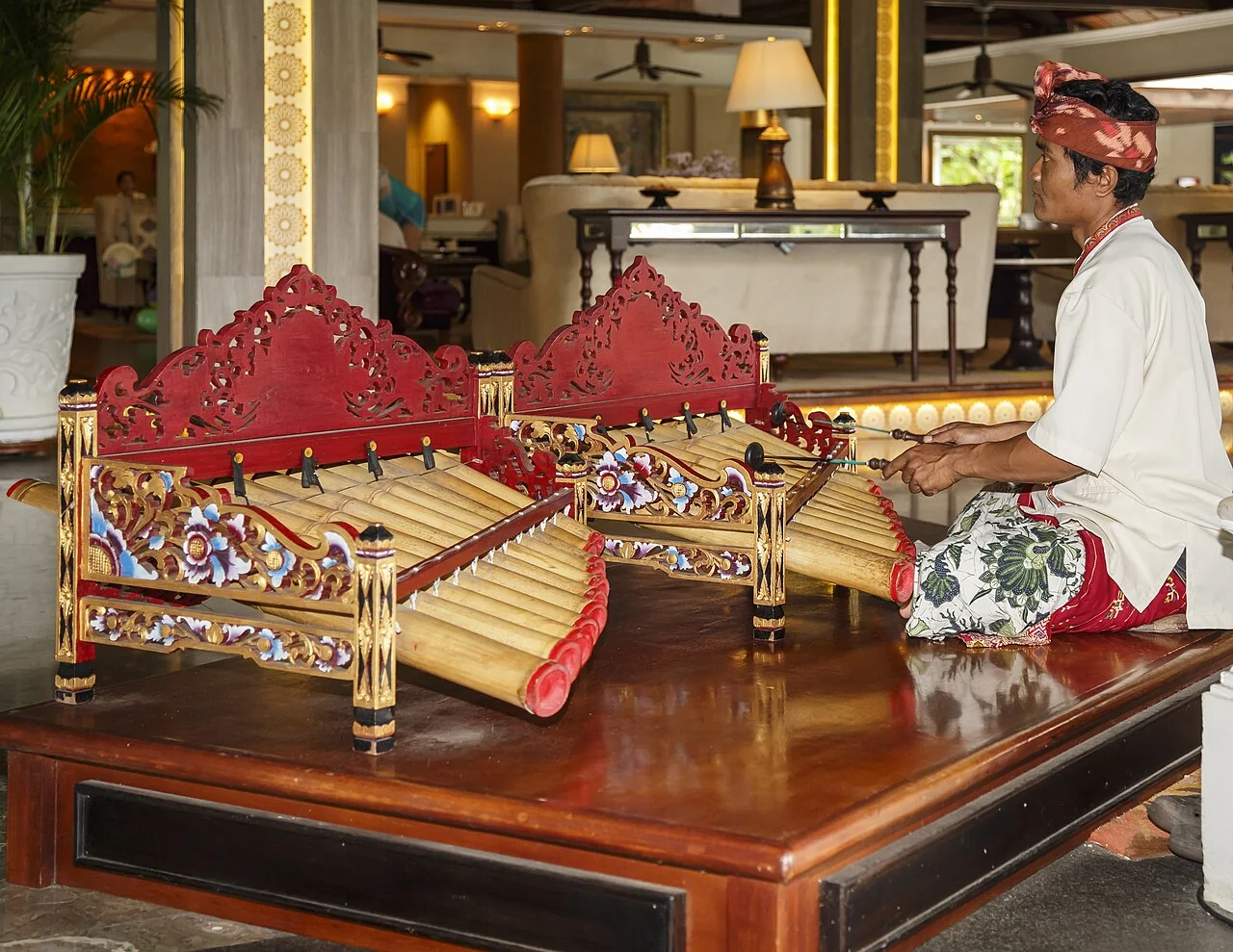 A man in traditional dress sits on a wooden platform playing a bamboo xylophone with mallets.
