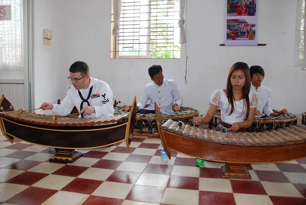 Musicians play wooden xylophones and circular sets of gongs indoors.