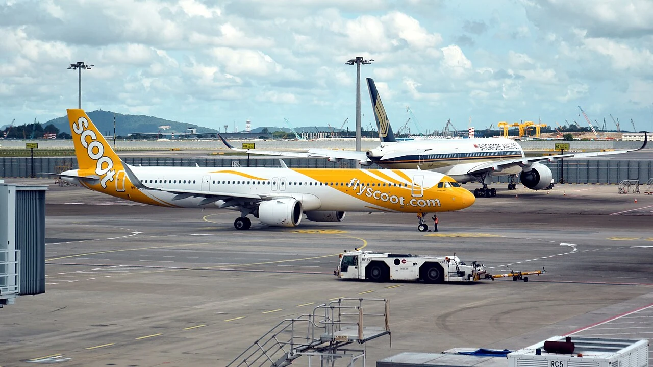 A low white pushback tractor pulls a long yellow towbar across the apron.