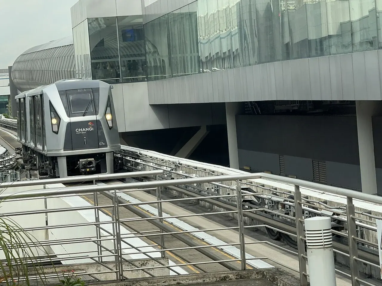 A silver airport train approaches along raised tracks beside a terminal building.